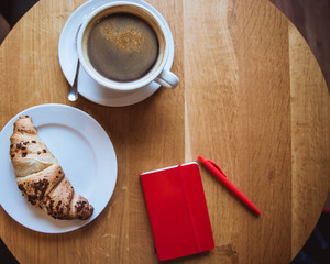 A red note and red pen is on the table in a cafe, a cup of coffee and a croissant