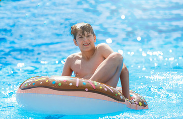 Cute child boy on funny inflatable donut float ring in swimming pool. Teenager learning to swim, has fun in outdoor pool at tropic resort. Water toys for kids. Healthy sport activity, vacation 