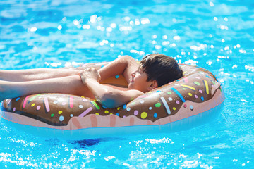 Cute child boy on funny inflatable donut float ring in swimming pool. Teenager learning to swim, has fun in outdoor pool at tropic resort. Water toys for kids. Healthy sport activity, vacation 