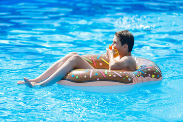 Cute child boy on funny inflatable donut float ring in swimming pool with oranges. Teenager learning to swim, have fun in outdoor pool at resort. Water toys for kids. Healthy sport activity, vacation 