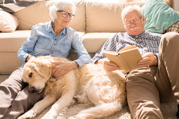 Portrait of happy senior couple cuddling with pet dog and reading books  sitting on floor at home ...