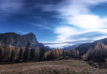 Naklejka na ściany i meble Mountains at beautiful night in autumn in Dolomites, Italy. Landscape with trees in alpine mountain valley, meadow, sky with clouds, moonlight, stars. Scenery with forest and rocks. Space 