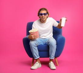 Emotional man with 3D glasses, popcorn and beverage sitting in armchair during cinema show on color...