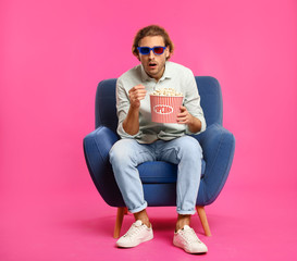 Emotional man with 3D glasses and popcorn sitting in armchair during cinema show on color background