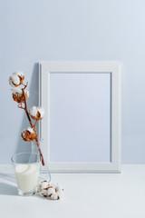 Mock up white frame, dry cotton twigs in vase and glass of milck on book shelf or desk. White-blue colors. Minimalistic concept.