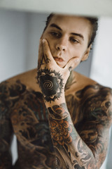 a young handsome tattooed man in the morning shaves in front of a mirror, a razor in his hands, a...