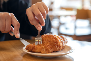 Closeup image of hands cutting a piece of croissant by knife and fork for breakfast on wooden table