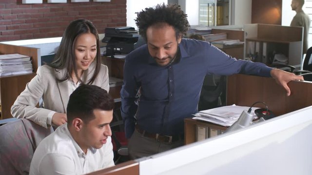 Group of three office workers of different ethnicities discussing work, one of them sitting at desk and the other two standing near him