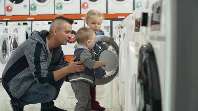 Cute little girl and her baby brother opening the door of washing machine and looking into it with dad while shopping together in home appliance store
