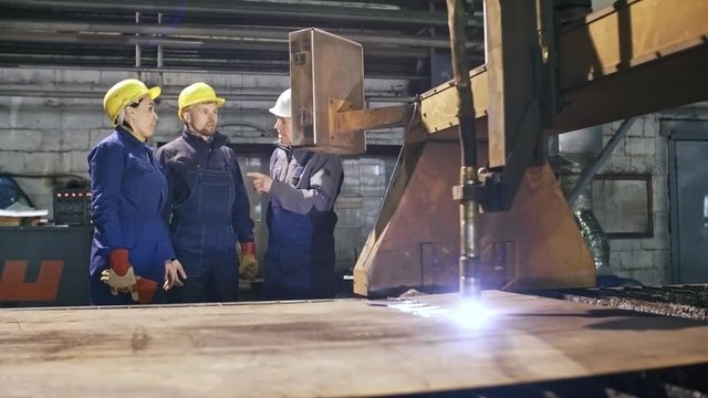 Medium shot of female and male engineers in hard hats and uniforms talking and pointing at control panel of metal laser machine cutting sheet