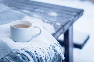 Winter concept, wooden table covered with snow in the backyard or park, cozy blanket and a cup of hot tea outdoors, minimal style, copyspace