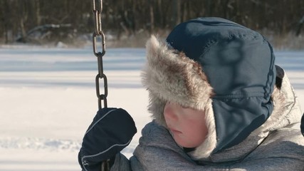Kid swaying on swing. Cute little boy (toddler, two year old) in hat with fur trim and earflaps.  Winter, snow, cold weather, outdoor activity