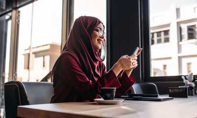 Woman in hijab sitting at cafe using mobile phone - obrazy, fototapety, plakaty