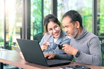 Fototapeta na wymiar Attractive mature asian man with white stylish short beard looking at laptop computer with teenage eye glasses hipster woman in cafe. Teaching internet online or wifi technology in older man concept.