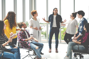 Attractive young multiethnic group talking at relax zone in coffee break time at office. Young asian hipster man sharing lifestyle idea to staff friends with fun and happy in happy workplace concept.