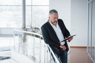 Big windows provide excellent lighting. Photo of senior businessman in the spacious room with plants behind. Holding and reading documents