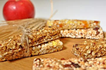 Side view close up of sweet slices of healthy cereal bars with dried wheat and fruits on brown wooden table and red apple background