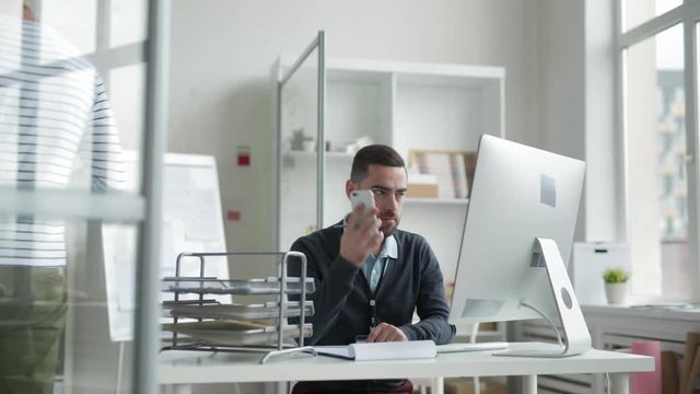 Tracking right of businessman with beard sitting at desk, writing and then answering telephone call, while his colleague passing with papers in his hands