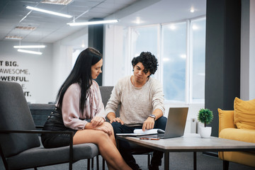 Fototapeta premium Sits on a grey chairs. Curly guy and brunette girl discusses the details of the contract in modern office