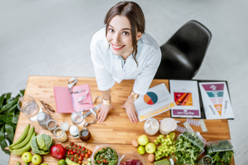 Portrait of a young woman nutritionist in medical uniform standing near the table full of various healthy products indoors