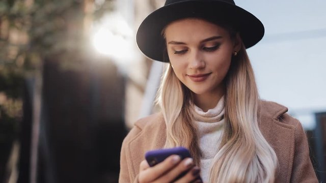 Attractive young woman wearing a coat and hat is standing in autumn street and looking at her smartphone, smiling. Social media and street fashion concept
