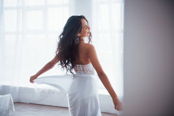 From behind. Beautiful woman in white dress stands in white room with daylight through the windows