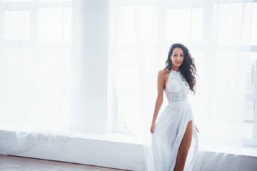 Fototapeta premium Looking straight into the camera. Beautiful woman in white dress stands in white room with daylight through the windows