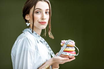 Portrait of a sad woman nutritionist in medical gown with donuts and tape measure on the green background. Unhealthy eating and adiposity concept