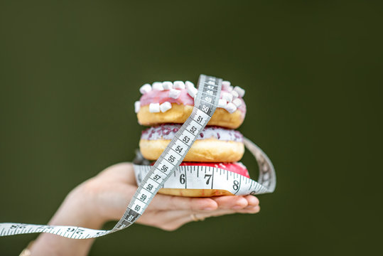 Three sweet donuts covered with measurement tape on the hand on the green background. Unhealthy eating and adiposity concept