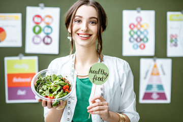 Portrait of a woman dietitian in medical gown standing with salad on the green wall background with...