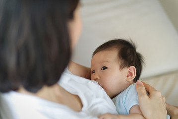 Young beautiful Asian mother sitting and holding her newborn child while breastfeeding her child on sofa. She is looking at her baby with eyes contact. Mother's breastfeeding concept.