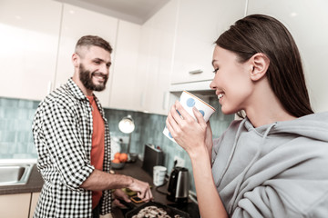 Smiling woman drinking tea while watching her man cooking