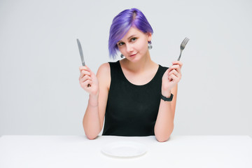 Fork, plate, knife - Concept portrait of a beautiful girl with purple hair on a white background sitting at a table with dining tools.