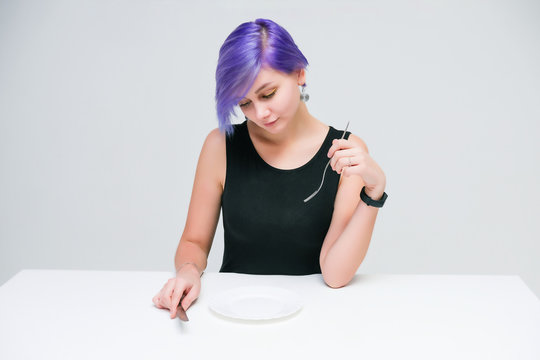 Fork, plate, knife - Concept portrait of a beautiful girl with purple hair on a white background sitting at a table with dining tools.
