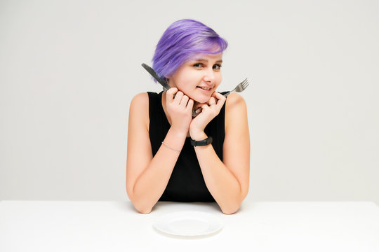 Fork, plate, knife - Concept portrait of a beautiful girl with purple hair on a white background sitting at a table with dining tools.