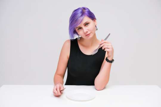 Fork, plate, knife - Concept portrait of a beautiful girl with purple hair on a white background sitting at a table with dining tools.