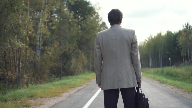 An elderly businessman in a jacket with a briefcase is standing on the road, and looking into the distance. Back view. Unknown feauture