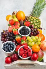 Fototapeta na wymiar Healthy fruits in a wooden tray, strawberries raspberries oranges plums apples kiwis grapes blueberries mango persimmon pineapple on the white table, copy space for text, selective focus