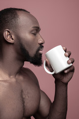 African man with white cup of tea, isolated on pink studio background. Close up portrait in minimalism style of a young naked happy afro man