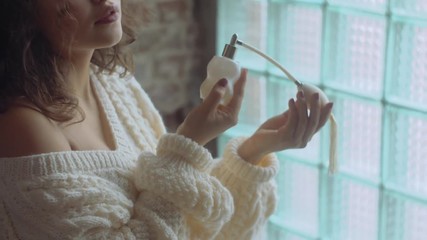Close up: Young beautiful confident fashionable woman using, holding luxury perfume in vintage white glass bottle with pump. Model wearing warm winter knitted sweater - Powered by Adobe