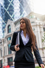 Junge Geschäftsfrau im Anzug und mit Laptop in der Hand steht vor modernen Bürogebäuden in der Stadt