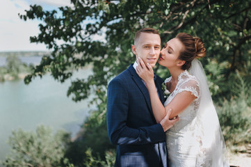 Beautiful newlyweds cuddle in nature, near the green tree. Cute brunette bride in a lace dress hugs and kisses on the cheek of a stylish groom in a suit and bow tie. Wedding photography.