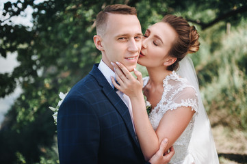 Beautiful newlyweds cuddle in nature, near the green tree. Cute brunette bride in a lace dress hugs and kisses on the cheek of a stylish groom in a suit and bow tie. Wedding photography. Close-up.