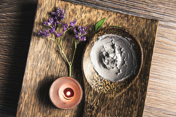 Natural gray mud mask cream on wooden spoon with candle burning and flower branch as decoration on wooden tray on table. Flat lay view, copy space. Cozy warm vintage look.