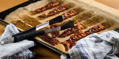 Knusprig gebratene Bacon Speck Scheiben aus dem Ofen auf Backblech mit Backpapier mit Geschirrtuch und Zange in Küche