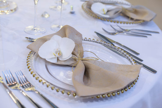 Fine dining table setting featuring transparent plates, beige linen napkin with natural orchid and golden decorations and silverware in the order of use, ready for guests at a formal event or wedding