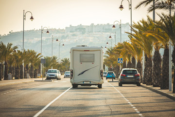 Motorhome arriving in summer paradise in street full of coconut palms and palm trees in beach town. Auto Caravan on the street seen from behind traveling from summer holidays
