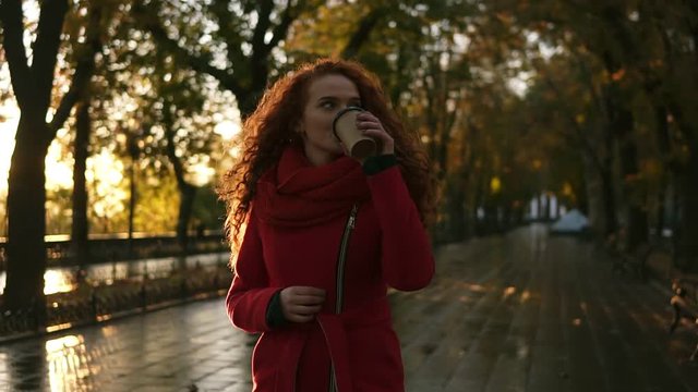 Red haired woman walking on a city park. Cold weather, she wears red colored coat and knitted scarf. Long haired beautiful, smiling girl drinks coffee from "to go" cup. Lens flares, golden sunlight
