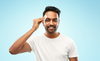 Fototapeta premium grooming, skin care and people concept - smiling young indian man cleaning his face with cotton pad over blue background