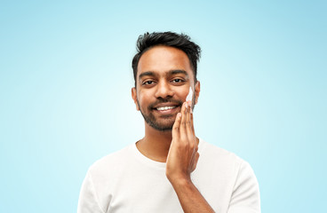 grooming, skin care and people concept - smiling young indian man applying cream to face over blue background